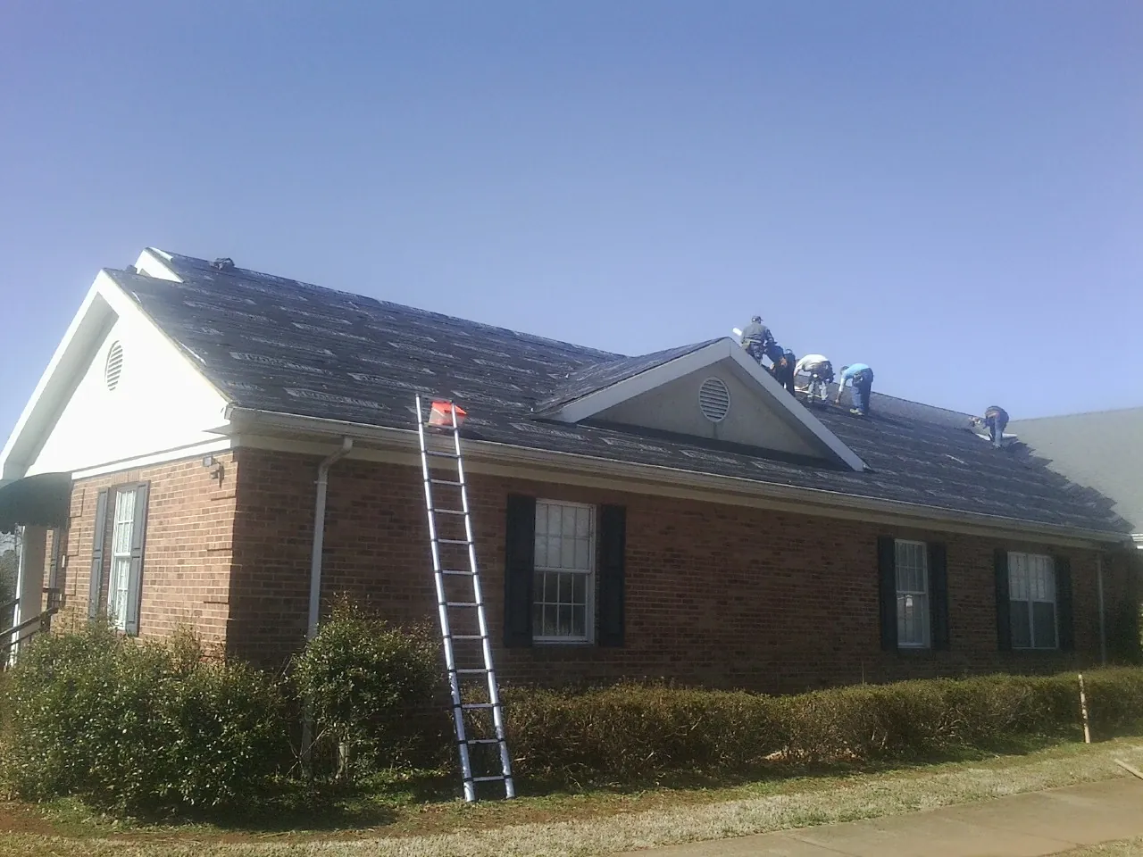 Crew tearing shingles off a brick ranch roof with a ladder set — Emergency Roof Repair work by our crew in Oak City